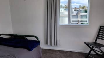 Quarto com cama, uma cadeira, janela com cortinas e vista para uma cerca e edifícios lá fora.
