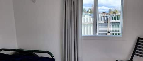 A bedroom with a bed, a chair, a window with curtains, and a view of a fence and buildings outside.