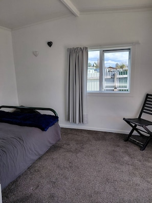 A bedroom with a bed, a chair, a window with curtains, and a view of a fence and buildings outside.