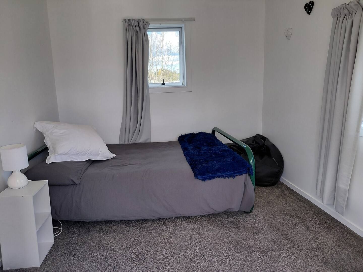 A bedroom with a bed, a nightstand, a lamp, a window with curtains, and a view of the outdoors.