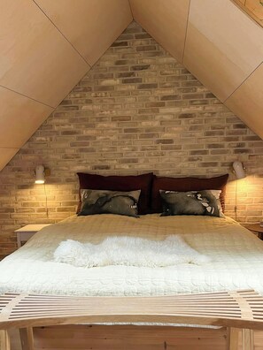 Ein Schlafzimmer mit schrägem Dach, einer Backsteinwand, einem Bett mit weißer Bettdecke und zwei Nachttischlampen.
