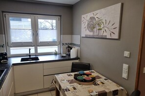 A compact kitchen with a window, a sink, a dining table with a coffee-themed tablecloth, and a wall-mounted painting. - Zugspitze 112 including sauna (Garmisch-Partenkirchen)