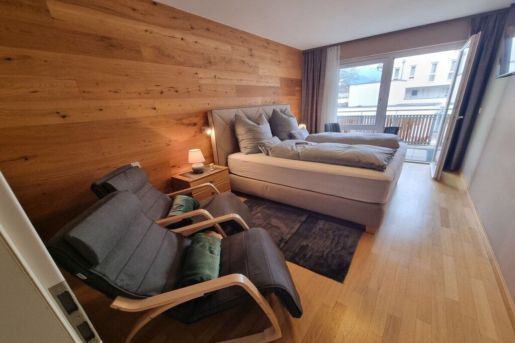 Une chambre d’hôtel avec un mur en bois, un lit avec une literie grise, une petite table de chevet avec une lampe, une chaise et une vue sur l’extérieur.