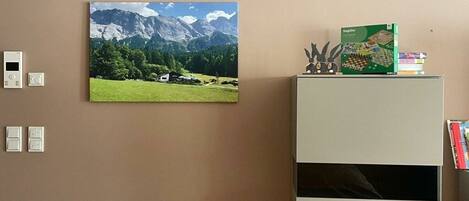 A room with a mountain landscape painting, a white cabinet with a book on top, and a thermostat on the wall.