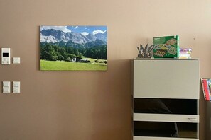 A room with a mountain landscape painting, a white cabinet with a book on top, and a thermostat on the wall. - Zugspitze 112 including sauna (Garmisch-Partenkirchen)