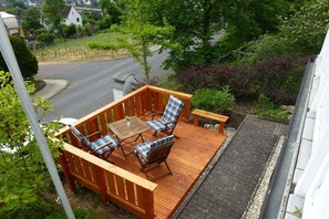 A wooden deck with a table and chairs, surrounded by greenery and a view of a road. - Holiday apartment with 1 bedroom (Treis-Karden)