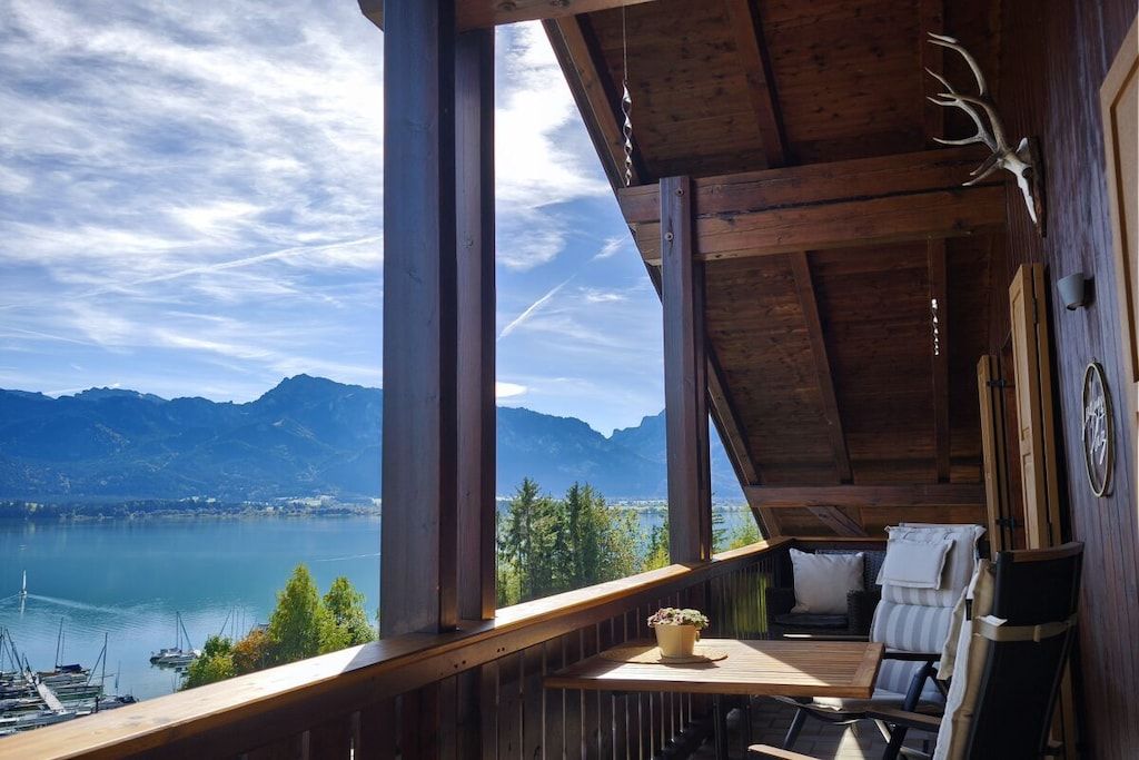 A wooden deck with a table and chairs, overlooking a lake and mountains.