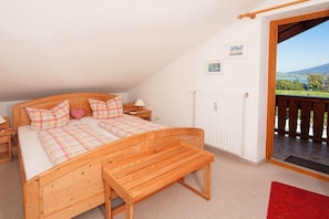 A bedroom with a wooden bed, a bench, a nightstand, a lamp, and a view of a balcony with a door. - Holiday apartment with 1 bedroom (Rieden am Forggensee)
