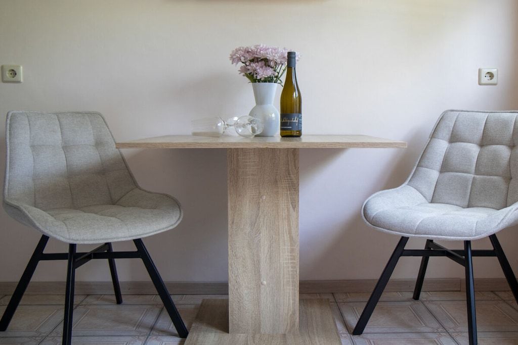 A room with two grey upholstered chairs, a wooden table, a vase with pink flowers, and a bottle of wine.