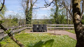 A fenced outdoor area with a table and chairs, surrounded by trees and a grassy field with white flowers.