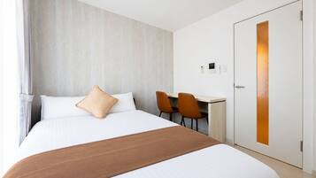 A hotel room with a bed, a desk, two chairs, a door, and a wall-mounted control panel.