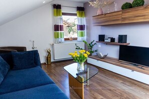 A modern living room with a sofa, a glass coffee table with a vase of flowers, a flat-screen TV, and wooden shelves. - Holiday apartment in Königheim with barbecue (Königheim)