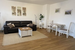 A modern living room with a black sofa, a white coffee table, and a dining area with a table and chairs. - Holiday apartment with 1 bedroom (Südheide\/ Hermannsburg)