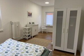A bedroom with a bed, a wardrobe, a radiator, a desk, a chair, and a window with blinds. - Holiday apartment with 1 bedroom (Südheide\/ Hermannsburg)