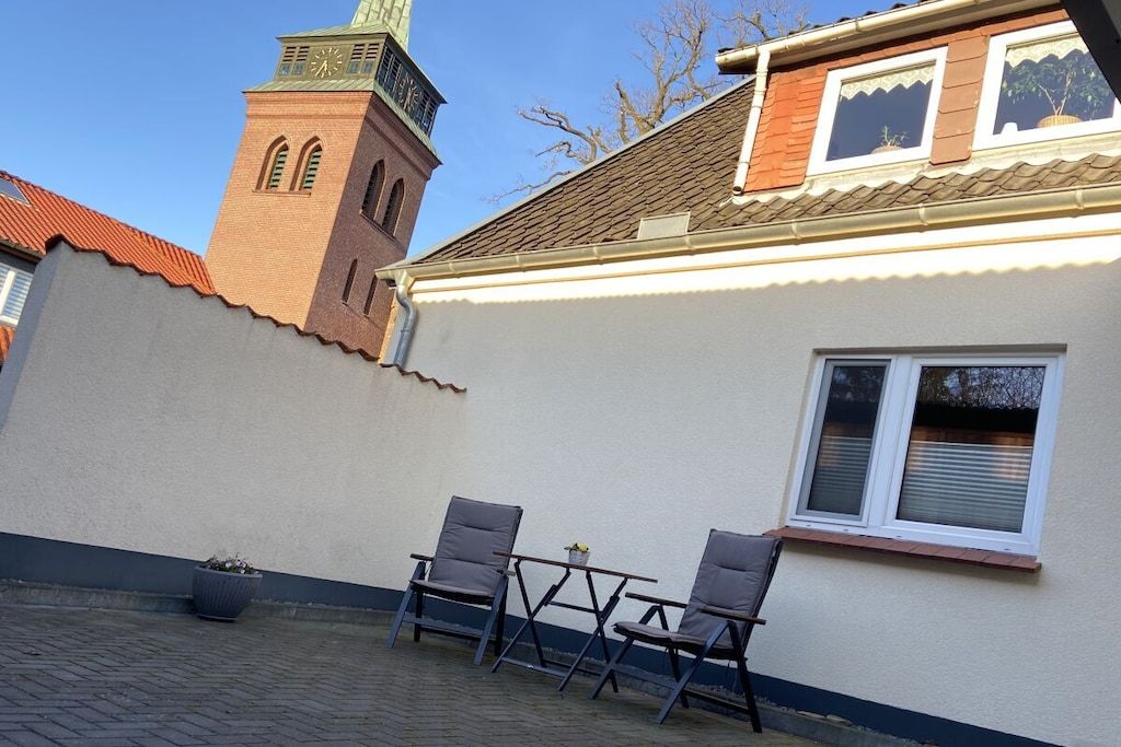 A building with a clock tower, a window with shutters, and a paved area with two chairs.