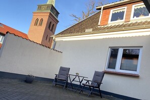 A building with a clock tower, a window with shutters, and a paved area with two chairs. - Holiday apartment with 1 bedroom (Südheide\/ Hermannsburg)