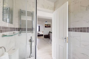 Un baño moderno con cabina de ducha de vidrio, repisa de vidrio y puerta blanca que da a una zona de estar con cama y un cuadro en la pared.