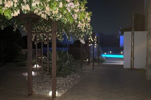 Un espace extérieur aménagé pour la nuit, avec une pergola en bois recouverte de fleurs, un chemin menant à une piscine et un bâtiment dont la façade, éclairée de lumière bleue, donne sur la piscine.