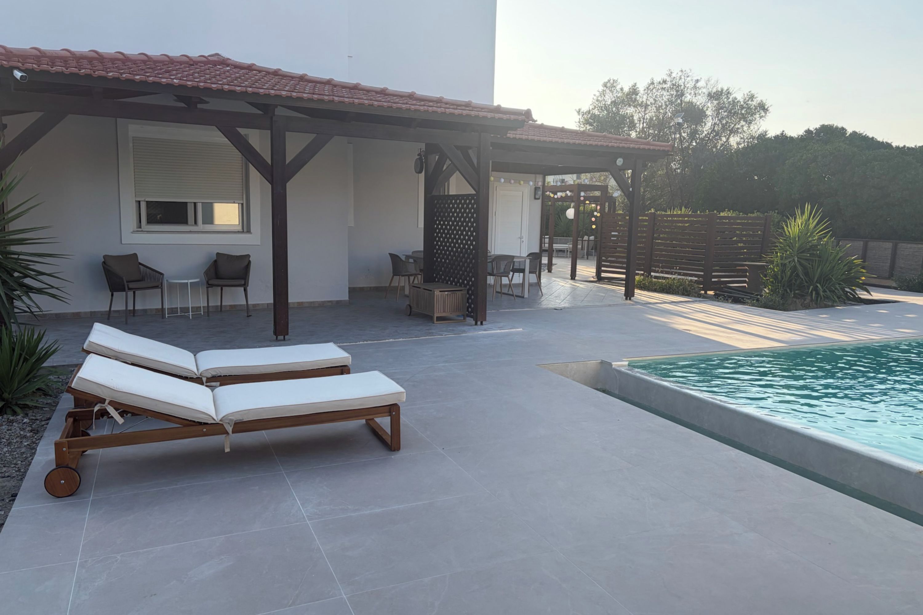 Un área junto a la piscina con sillones reclinables, una pérgola y un porche cubierto con asientos.