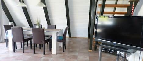Un área de comedor con una mesa y sillas, un televisor de pantalla plana sobre un soporte y una escalera con barandas de madera.