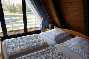 A room with two beds, a wooden ceiling, and a window with a view of a fence and trees. - Holiday home with 2 bedrooms (Butjadingen)