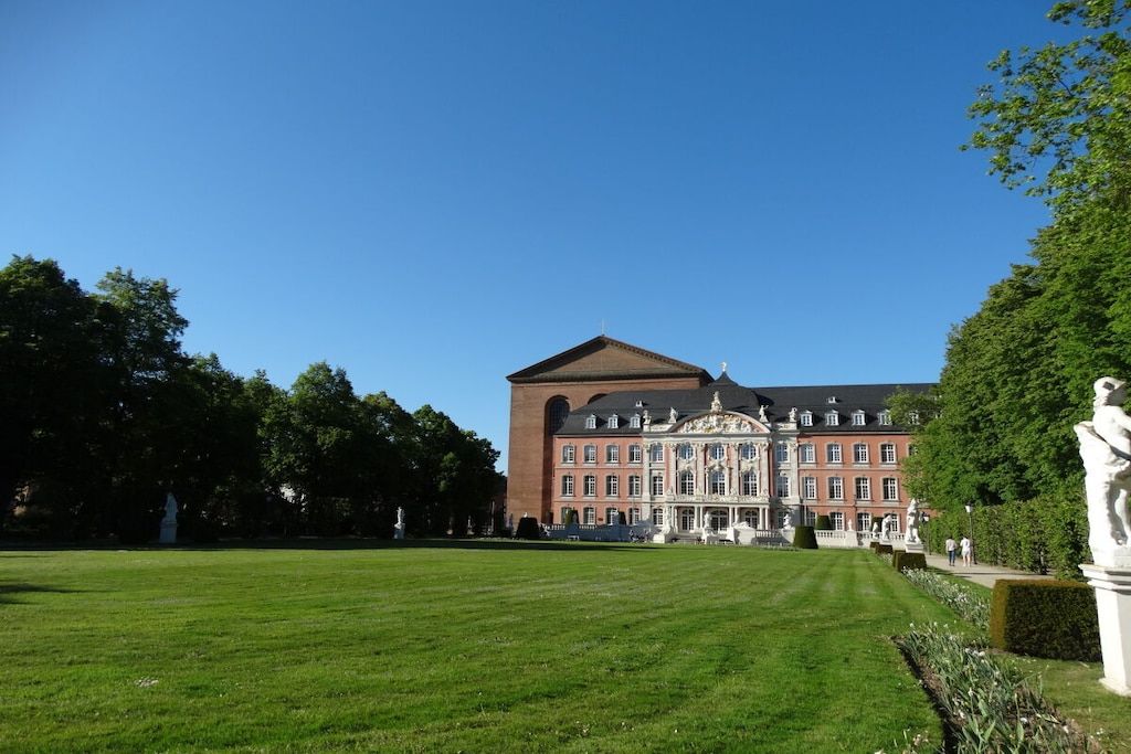 Ein großes, historisches Gebäude mit einer symmetrischen Fassade, einem zentralen Eingang mit einem verzierten Giebel und einem gepflegten Rasen davor.