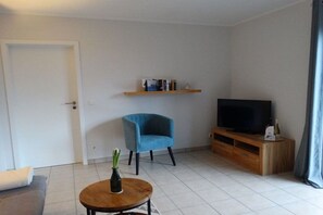 Een woonkamer met een bank, een rond bijzettafeltje met een vaas, een blauwe fauteuil, een televisie op een houten dressoir en een boekenkast.