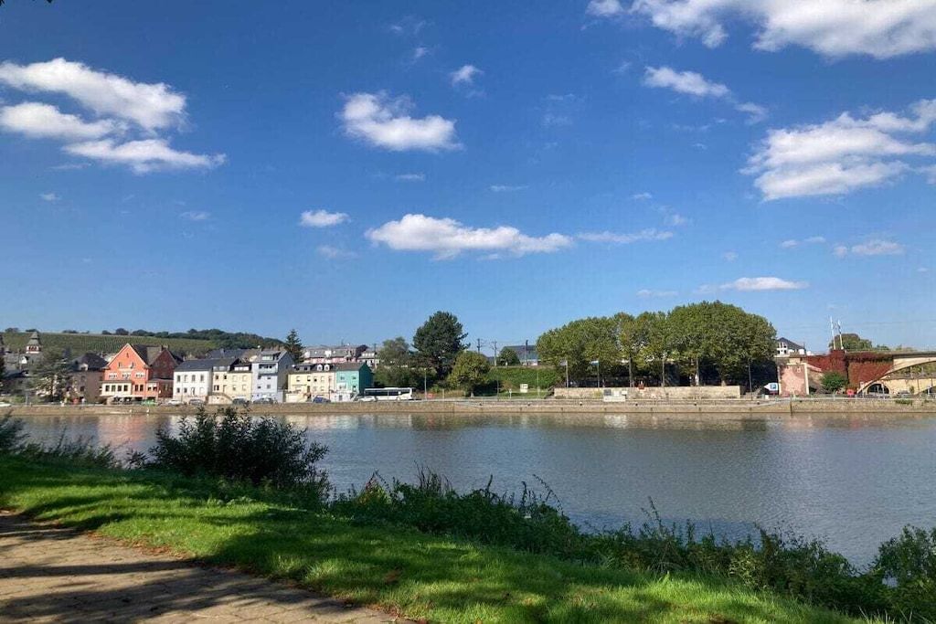 A riverside view with buildings along the water, a clear sky, and greenery.