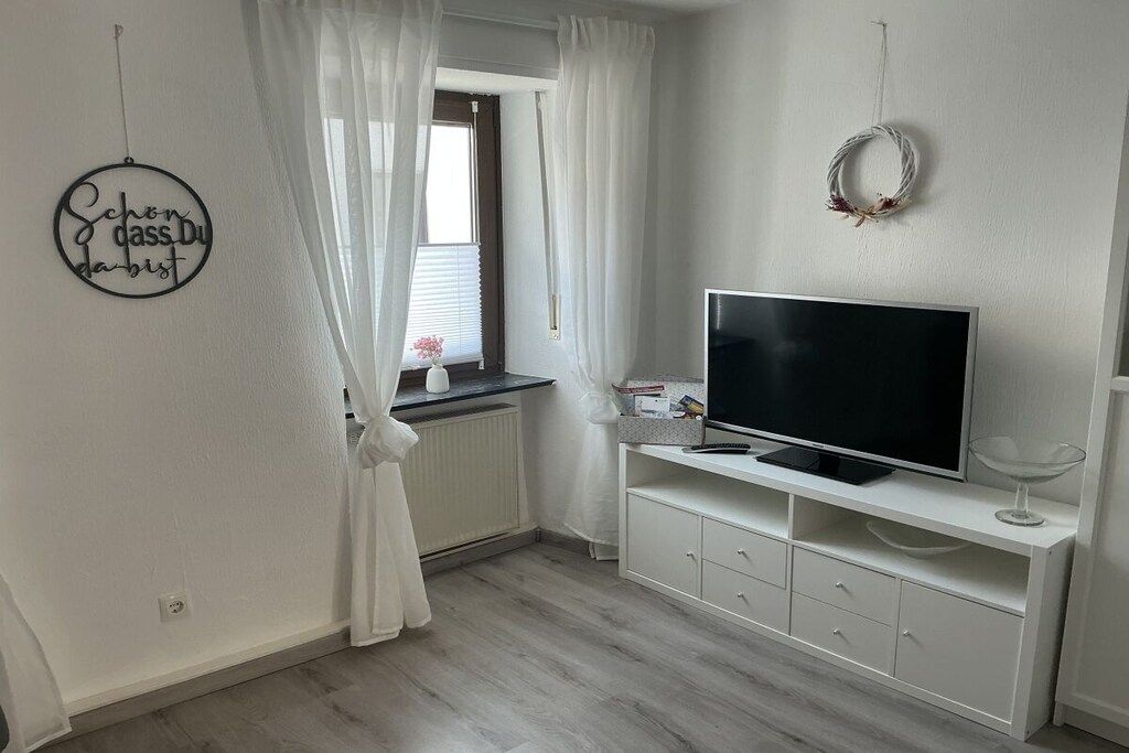 Ein Zimmer mit Fernseher auf einer weißen Kommode, einem Fenster mit weißen Vorhängen und einer Wanddekoration.