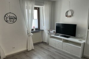 A room with a television on a white cabinet, a window with white curtains, and a wall decoration. - Holiday home with 2 bedrooms (Koblenz-Bassenheim)