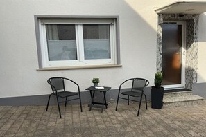 A small outdoor seating area with two chairs and a table, a window with blinds, and a door with a stone facade. - Holiday home with 2 bedrooms (Koblenz-Bassenheim)