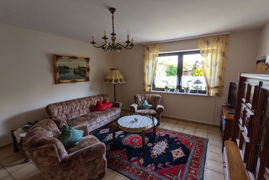 A living room with a sofa, armchairs, a coffee table, a chandelier, a painting, and a window with curtains.