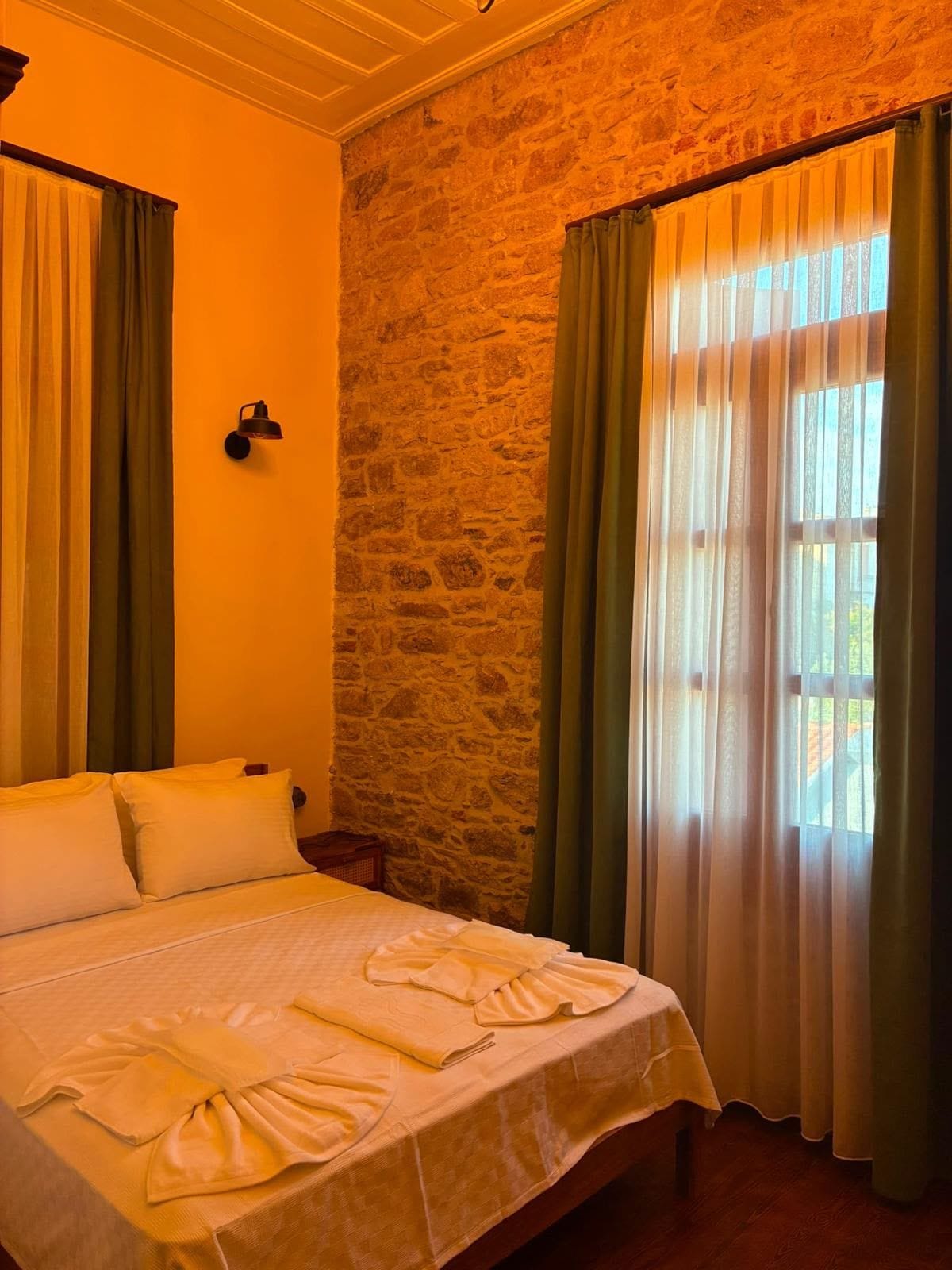 Habitación de hotel con pared de piedra, cama con ropa blanca y cortinas verdes.