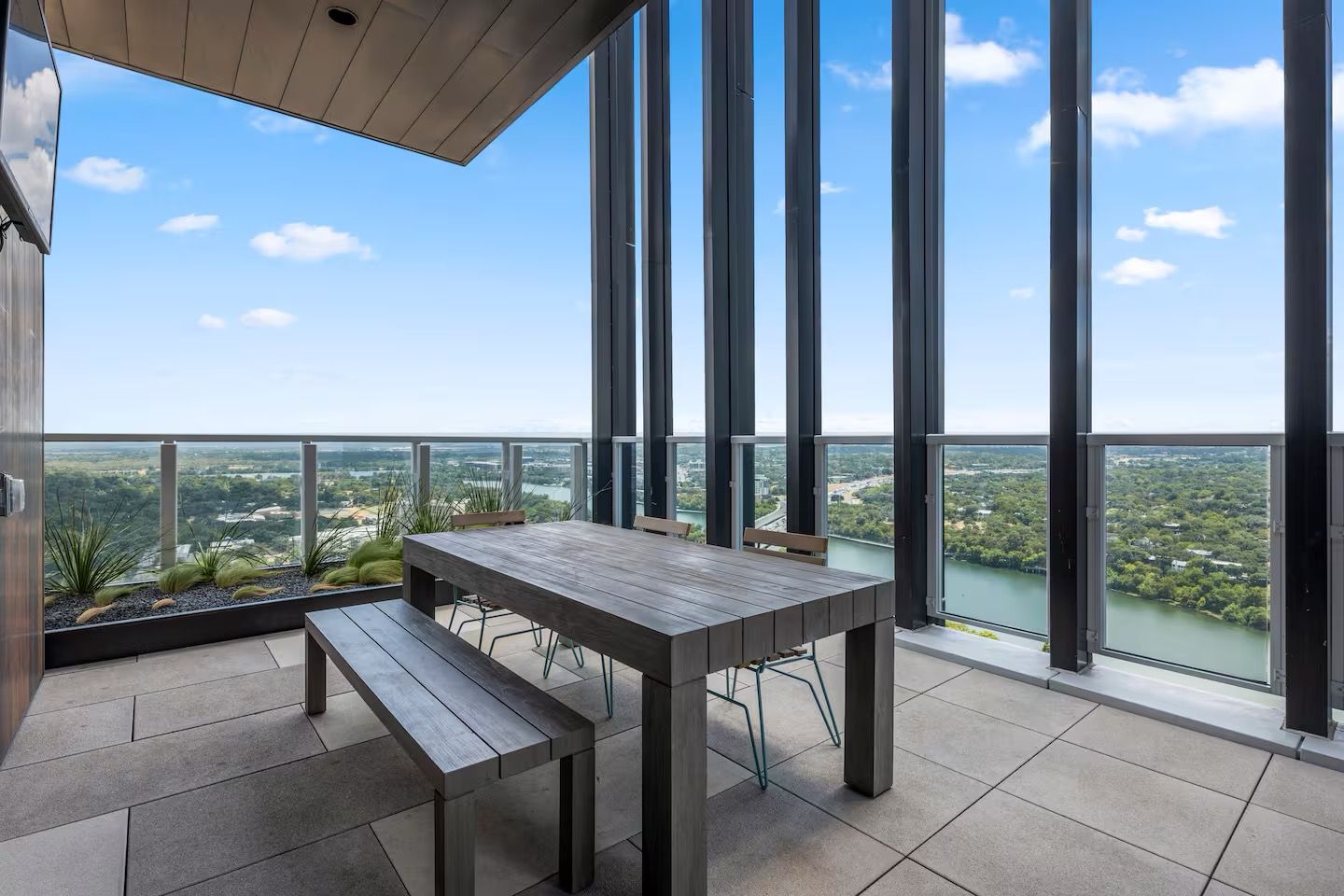 A balcony with a table and bench, large windows, and a view of a river and greenery.