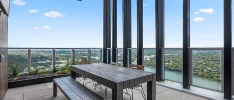 A balcony with a table and bench, large windows, and a view of a river and greenery.