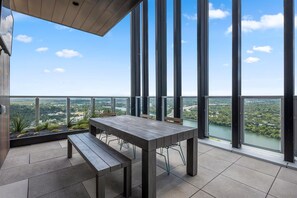 En balkon med bord og bænk, store vinduer samt udsigt over en flod og grøn vegetation.