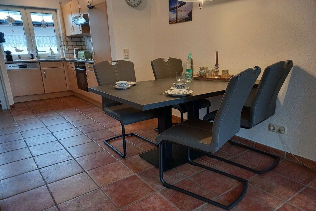 A dining area with a table and chairs, a kitchen in the background, and a tiled floor.