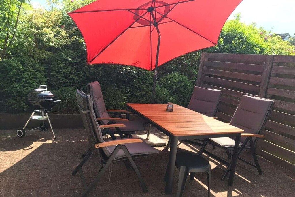 An outdoor patio with a wooden table and chairs, a red umbrella, and a barbecue grill.