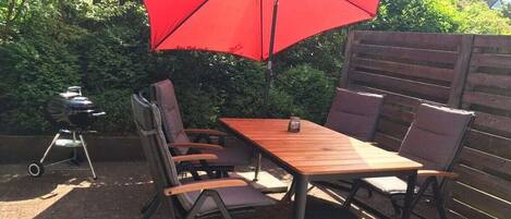 An outdoor patio with a wooden table and chairs, a red umbrella, and a barbecue grill.