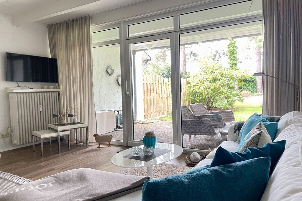 A modern living room with a flat-screen TV, a dining area with a table and chairs, and a large sliding glass door leading to a garden.
