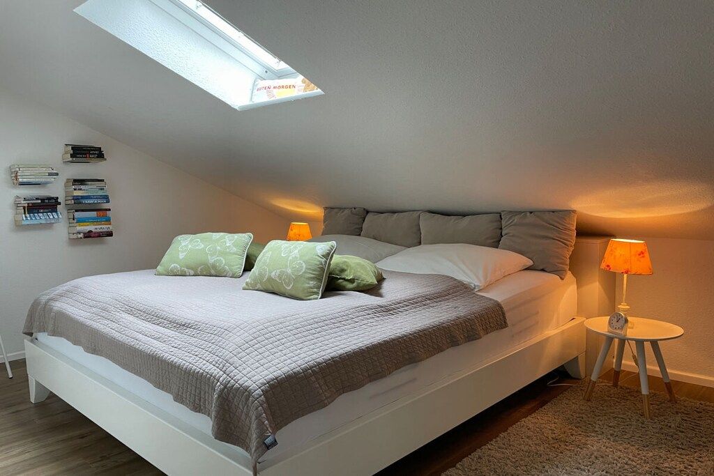 A bedroom with a sloped ceiling, a bed with a quilted bedspread, two pillows, a bedside lamp, and a small table with a clock.