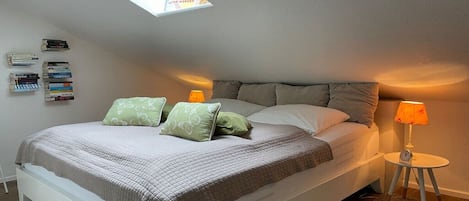 Un dormitorio con techo inclinado, cama con colcha acolchada, dos almohadas, una lámpara de noche y una mesita de noche con un reloj.