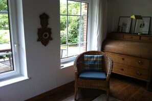 Une pièce avec un fauteuil en osier, une commode en bois et une fenêtre donnant sur un paysage verdoyant.