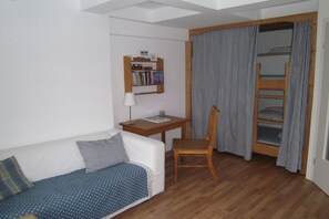 Une petite pièce avec un bureau en bois, un canapé blanc, une chaise, une bibliothèque et un lit superposé avec des rideaux.