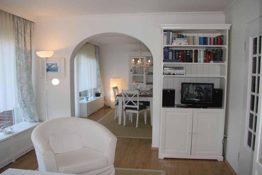 A living room with a white armchair, a dining area with a table and chairs, a bookshelf with books and a television, and a window with curtains.