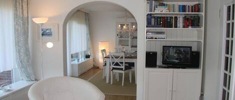A living room with a white armchair, a dining area with a table and chairs, a bookshelf with books and a television, and a window with curtains.