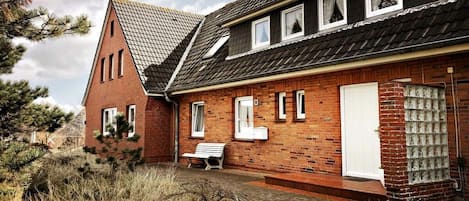 A two-story brick house with a sloped roof, a front porch with a bench, and a small garden area.