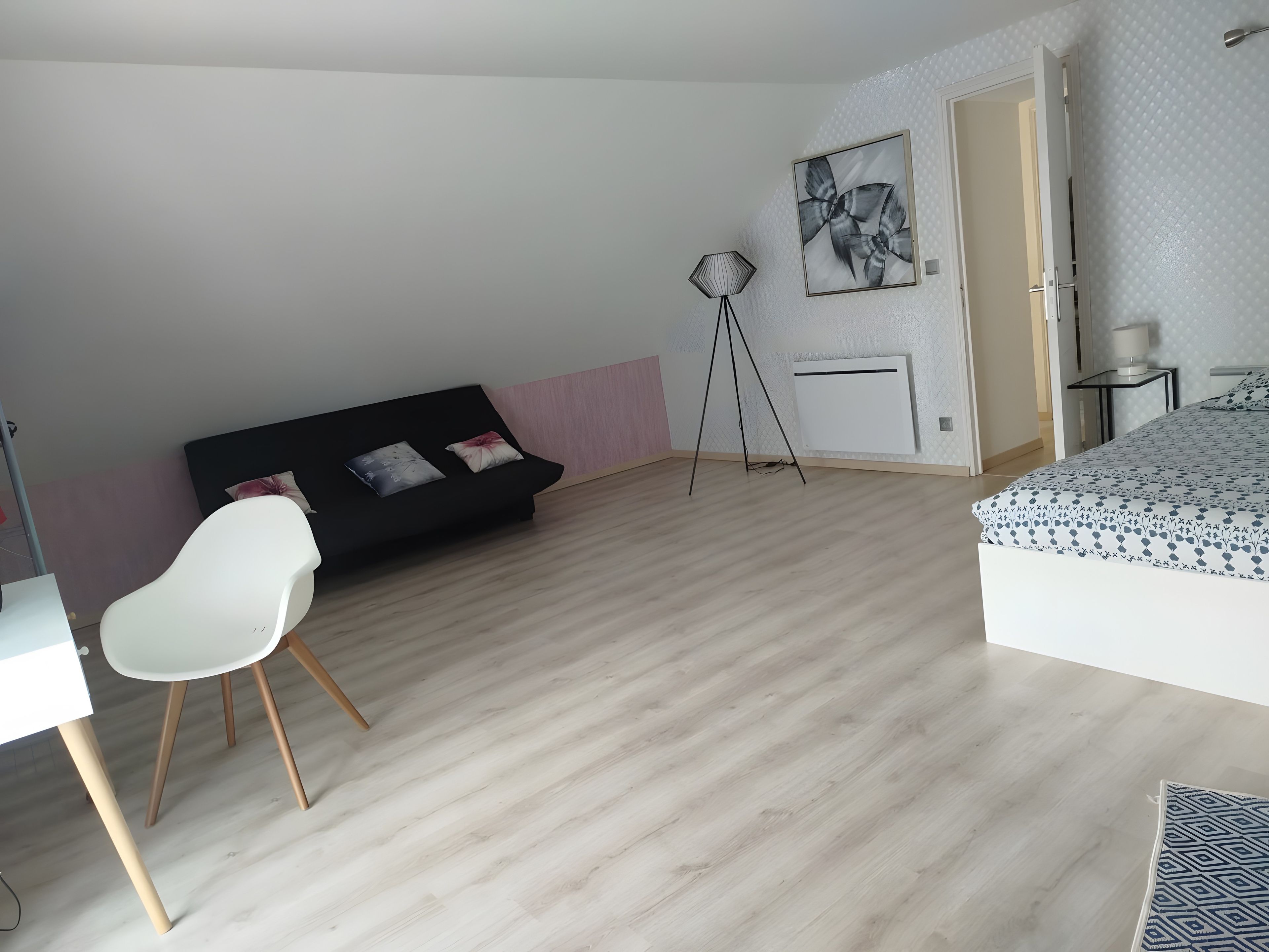 A room with a black sofa, a white chair, a bed with patterned bedding, a floor lamp, and a radiator.
