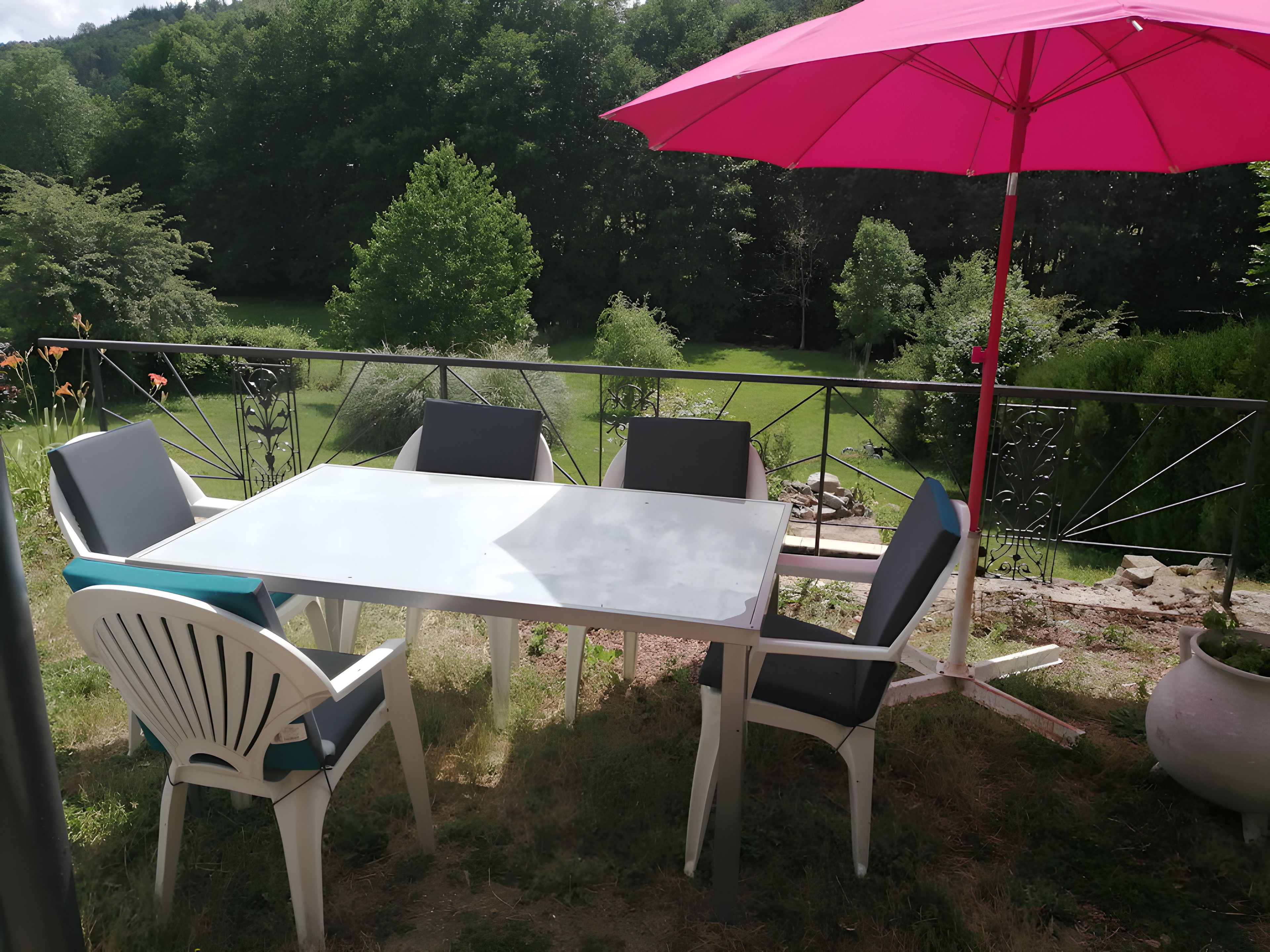 Une terrasse extérieure avec une table et des chaises, un parasol rouge et une vue sur un jardin et des arbres.