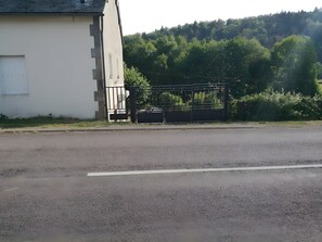 A paved road with a white line marking, a metal gate, and a building with a white facade. - Gîte Le Petit Buis: Mountain Views, Private Terraces & Wi-Fi (Chissey en Morvan)